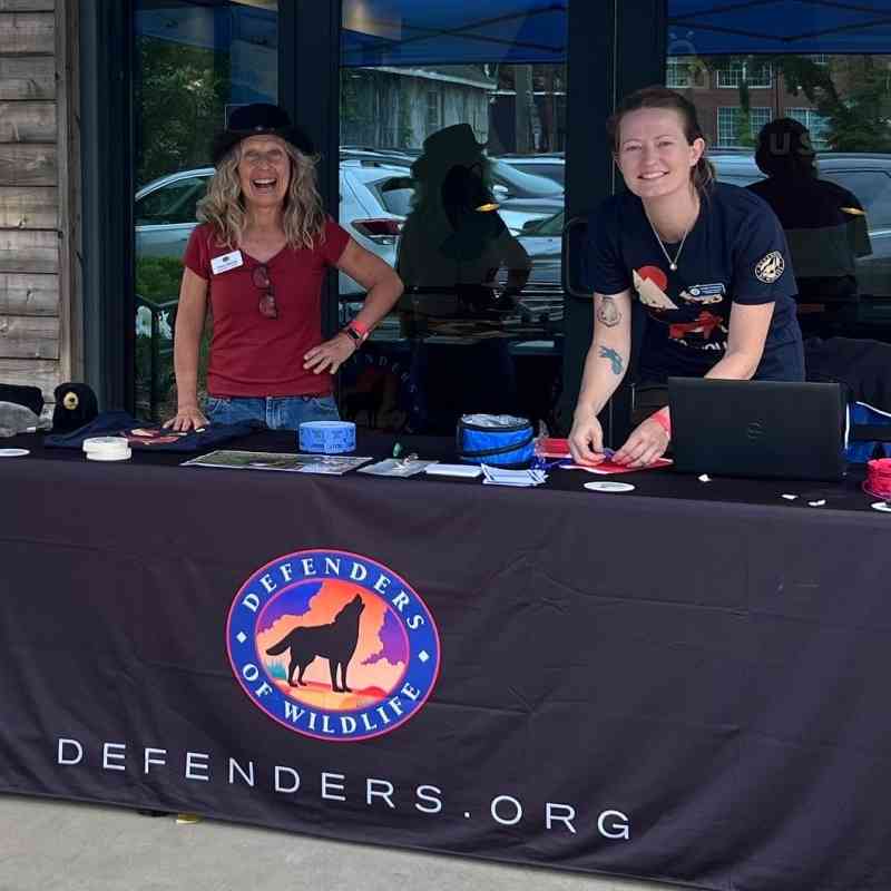 Tracy Davids and Heather Clarkson stand behind a Defenders of Wildlife table under a blue tent outside.